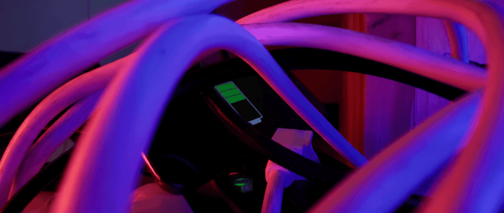 A close-up shot showing a charging battery icon on a black screen, partially obscured by several tangled, thick white cables under vibrant purple and red lighting.